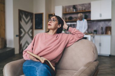 Genç, orta yaşlı, gözlüklü Asyalı bir kadın kanepeye oturuyor ve elleriyle başını çeviriyor ve evde rahat bir oturma odasında dinleniyor.