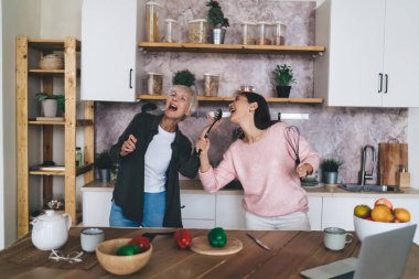 Evde birlikte yemek pişirirken mutfak gereçlerini mikrofon olarak kullanıp şarkı söyleyen çok ırklı kadın akrabalar.