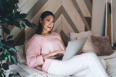 Pembe kazaklı, beyaz pantolonlu, rahat bir yatakta oturan, hafta sonu yatak odasında dinlenirken bilgisayar kullanan yaşlı, etnik bir kadın.