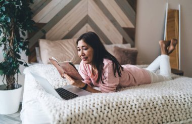 Orta yaşlı, gülümseyen ve yatakta dinlenen Asyalı bir kadın. Boş zamanlarında rahat yatak odasında defter okurken.