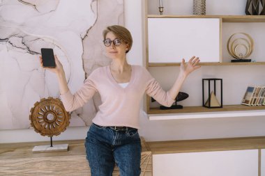 Gözlüklü, akıllı telefon ve kolları havada, evde çalışırken kameraya bakarken beyaz panoda fotokopi alanı gösteren sıradan kıyafetleri olan yetişkin bir iş kadını.