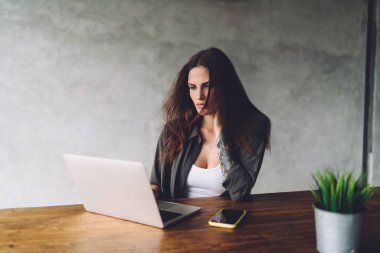 Düşünceli genç bayan serbest çalışan gündelik kıyafetlerle tahta masada dizüstü bilgisayarı ve akıllı telefonuyla uzaktan kumandalı bir proje üzerinde çalışıyor.