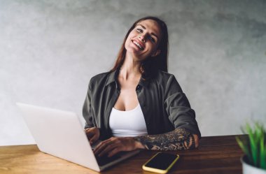 Kolunda dövmesi olan, gülümseyen, cep telefonuyla masada oturan ve kafedeki iş projesinde uzaktan çalışırken internet üzerinden yazan serbest çalışan bir kadın.