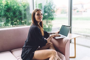 Kanepede oturup bilgisayarla iş projesi üzerinde çalışırken gülümseyen ve kameraya bakan genç bir kadının yan görüntüsü.