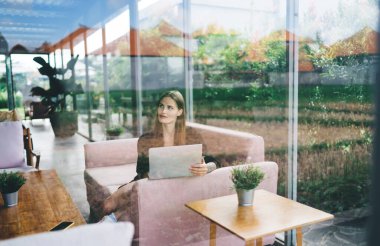 Uzun saçlı genç bayan girişimcinin penceresinden kanepeye oturup, gözlerini kaçırarak kafede dizüstü bilgisayar kullanarak uzak bir iş kurma girişiminde bulunuyor.