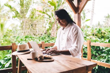 Yetişkin Asya 'lı erkek girişimcilerin günlük kıyafetleriyle dizüstü bilgisayarda çalışırken ve tahta masada kahve ve atıştırmalıklarla otururken ekrana bakarken yan görünümü