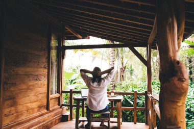 Dış ofisteki netbook üzerinde çalışırken terastan görülen ve Bali manzarasının tadını çıkaran sıradan kıyafetli yüzsüz Endonezyalı girişimcinin