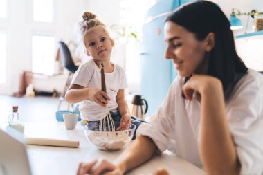 Bir kase un ve hamur işiyle masada oturan tatlı küçük kız dizüstü bilgisayarını kullanarak ve pasta tarifini okuyarak annesine bakarken...