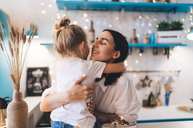 Mutlu genç anne gülümseyen ve sarılan kızı mavi mutfakta ve vazoda arpa pirinç bitkisi masaya yerleştirilmiş ve tatil hazırlıkları sırasında sıcak ışıklar açık