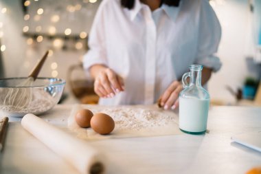 Beyaz elbiseli isimsiz bir kadın elinde un yumurtaları ve kâsenin yanında bir kavanoz sütle bayram için kağıt rulo pişiriyor.