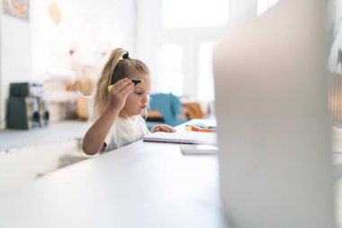 Beyaz tişörtlü, at kuyruklu, mutfak tezgahında elinde pastel kalemlerle oturan ve elinde sarı kalemle defterine bakan kızın yan görüntüsü.