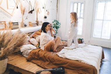 Beyaz gömlekli mutlu bir anne yatak odasında dinlenirken çıplak ayaklı kızın fotoğraflarını çekiyor.