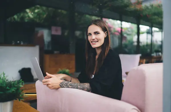 stock image Side view of young smiling female freelancer sitting in sofa near wooden table with green potted plants and looking at camera while working online