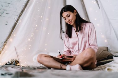 Genç Asyalı kadın evde dizüstü bilgisayar ve çelenklerle çadırda otururken deftere bir şeyler yazıyor. Eğlence ve eğlence anlayışı. Pijama giyen çekici bir kız. Noel ve Yeni Yıl Zamanı