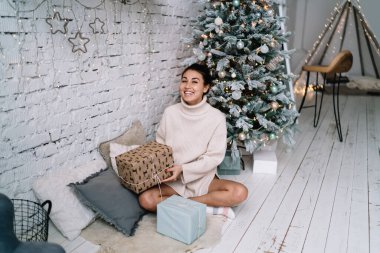 Genç gülümseyen Asyalı kadın tüylü halıda oturuyor ve geniş bir evde Noel ağacının yanında hediye kutusu tutuyor. Noel ve yeni yıl zamanı. Milenyum neslinin çekici kızı. Yurt içi yaşam tarzı