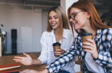 Gülümseyen, sıradan kıyafetlerle genç bayan öğrenciler masada sıcak içeceklerle otururken ve ödevlerini yaparken gülümsüyorlar.