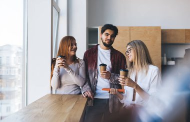 Bir bardak sıcak içecekle neşeli genç meslektaşlar pencerenin yanında dikilip modern ofisin bulanık önplanında gülüyorlar.