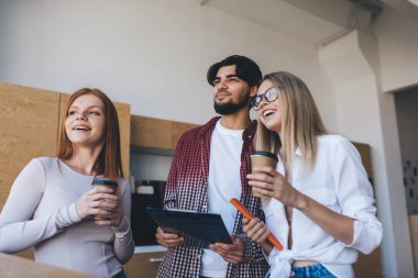 İş arkadaşlarından oluşan düşük açılı bir ekip, elinde not panosu olan erkek meslektaşı ve kahve fincanlarıyla gülümseyen kadın meslektaşları ofiste yan yana dururken...