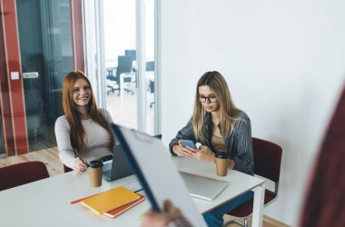 Neşeli bir kadın, meslektaşı ile oturmuş dizüstü bilgisayarlı, kahve fincanlarıyla akıllı telefon ekranına bakıyor. Proje sınıfında elinde pano olan tanınmayan bir erkek arkadaş sınıfındayken kameraya bakıyor.