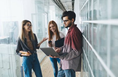 Ellerinde not defteri ve panosu olan genç kadın işçiler modern binanın cam duvarının yanında duruyor ve ekrana bakarken erkek meslektaşlarıyla bilgisayarla etkileşime geçiyorlar.