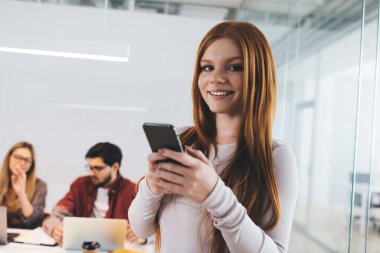 İş projesinde çalışırken cep telefonu kullanan ve kameraya bakan ve bilgisayarlı meslektaşlarına karşı duran memnun kadın ofis çalışanlarından.