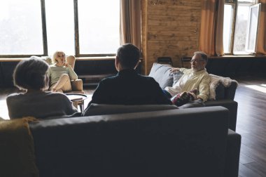 Modern oturma odasında büyük pencereli bir grup arkadaş haftasonu dinlenirken ve birlikte vakit geçirirken sohbet ediyorlar.