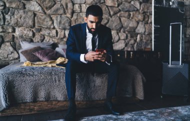 Şık takım elbiseli yakışıklı girişimci, ekranlara bakarken ve otel odasında dinlenirken mesajları ararken cep telefonunu karıştırıyor.