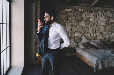 Zarif giyinmiş sakallı bir erkek pencerenin yanında dikilip, telefonla konuşurken yüzünü çeviriyor.