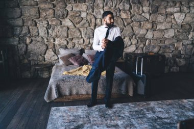 Yatak odasında dikilip ceketini çıkarırken, gece kıyafetlerini giyerken otel odasında dinlenen genç bir erkek.