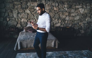 Etnik sakallı bir erkeğin işe gitmeden önce resmi kıyafetlerini giyip cep telefonuyla sohbet etmesi ve mesajlaşması.