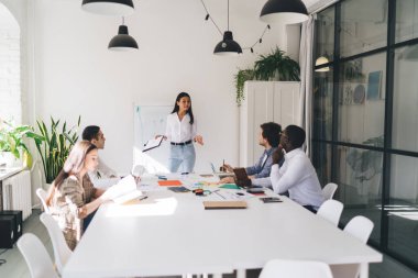 Cam duvarın yanındaki beyaz masada oturan çok ırklı çalışanlardan oluşan bir grup. Kadın meslektaşı ise elleri açık bir şekilde raporunu sunuyor.