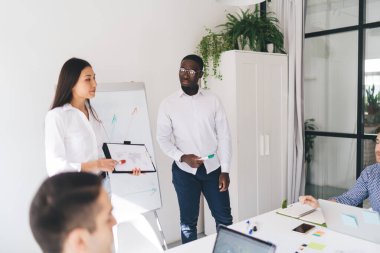 Modern yönetim kurulunda toplanan çok ırklı iş arkadaşları, Afrika kökenli Amerikalı meslektaşlarıyla yeni iş planlarını açıklarken kadın konuşmacının panoya işaret etmesini dinliyor.