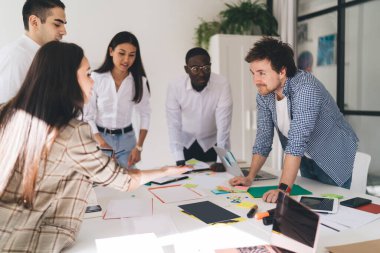 Çok ırklı tasarımcılardan oluşan bir grup yeni yaratıcı proje üzerinde çalışan araçlar ve belgelerle masanın etrafında toplanıyor ve fikirleri paylaşıyorlar.