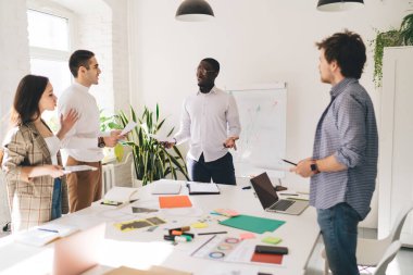 Modern ofiste birlikte çalışırken projeye başlama detaylarını tartışırken elinde kağıt dizüstü bilgisayar işaretleyicileri ve not defteriyle toplantı masasının yanında duran çok ırklı iş arkadaşlarının yan görünümü