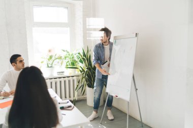 Beyaz panonun yanında elinde kalem ve kağıtlarla duran erkek sunucunun cam tarafından bakıldığında, ofiste katılımcılara proje detaylarını açıklarken