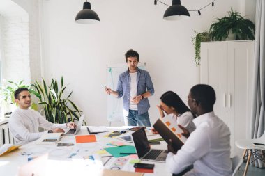 Bir grup çok ırklı iş arkadaşı dizüstü bilgisayarları ve gazeteleriyle masada toplanırken, erkek çalışanlar yazı tahtası önünde projeden bahsediyorlar.