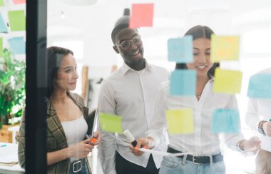 Şeffaf duvardaki parlak çıkartmalardan ve genç yaratıcı çeşitlilikteki insanlardan yeni plan fikirleri üzerinde çalışırken kağıtlarla çalışmalarına bakın