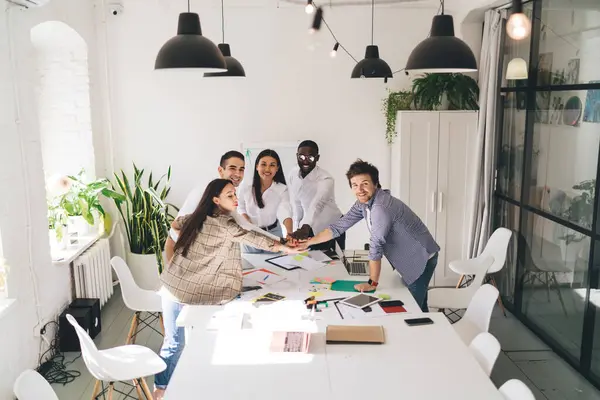 stock image High angle view of group of multiracial team workers gathering around table with papers laptop and tablet while looking at camera and joining hands in stack together during working on project in office