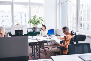 Modern ofisteki iş arkadaşlarının yanında proje üzerinde çalışırken masa başında oturmuş gündelik giysiler içinde konsantre olmuş bir erkek girişimci.