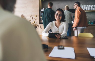 Gülümseyen kadın çalışan, arka planda erkek iş arkadaşlarıyla yaratıcı ofisteki ortakla proje tartışırken dijital tablet kullanarak masada oturuyor.