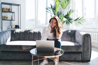 Odaklanmış İspanyol kadın serbest yazar akıllı telefonuyla rahat koltukta oturuyor ve boş zamanlarında bilgisayar ekranına bakarken ve evde bilgi ararken konuşuyor.