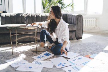 Asyalı kadın girişimcinin yan görüntüsü. Uzun saçlı, günlük kıyafetleri içinde. Oturma odasındaki masaya koyulan netbook 'a kağıtlar yazarken yerde oturuyor.