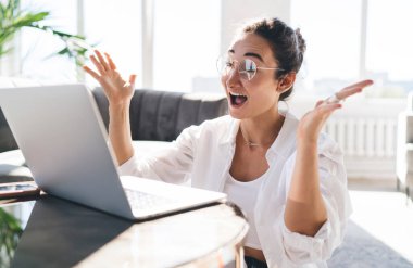 Beyaz elbiseli, gözlüklü, elleri keyifli bir şekilde masada oturmuş dizüstü bilgisayar ekranına bakan mutlu etnik kadın serbest yazar.