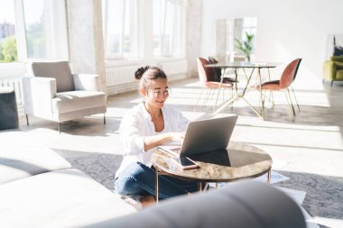 Netbook 'ta yazan ve gülümseyen, dağınık kağıtlarla masada oturan ve evdeki oturma odasında uzaktan kumandalı iş projesinde çalışan tam boy kadın serbest yazar.