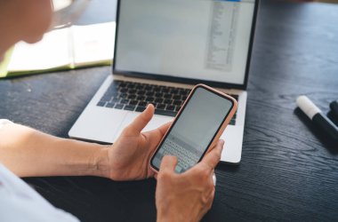 Yüksek açılı, tanınmayan serbest çalışan, ahşap masada dizüstü bilgisayarla oturuyor ve cep telefonuna kopyalama alanı sohbeti ve mesajlaşması ile bakıyor.