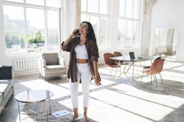 Evde boş zamanlarında masanın yanında durmuş cep telefonuyla konuşurken gülümseyen ve başka tarafa bakan pozitif genç bir kadın.