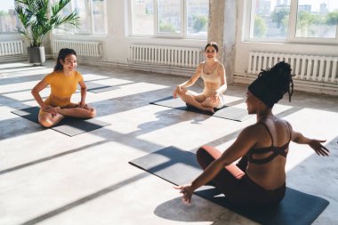 Aktif giyimli bir grup çok ırklı bayan sporcu güneşli spor salonlarında minderlerin üzerinde oturuyor ve sabah yoga sırasında nilüfer pozu alıştırması yapıyor.