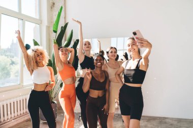 Spor yaptıktan sonra spor salonunun penceresinde beklerken, kolay giyimli ve selfie pozu veren heyecanlı kadın sporcular.