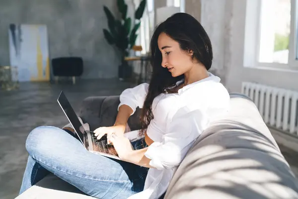 stock image Side view of positive Asian female worker wearing casual outfit messaging on netbook while sitting om comfortable sofa in creative workspace during break