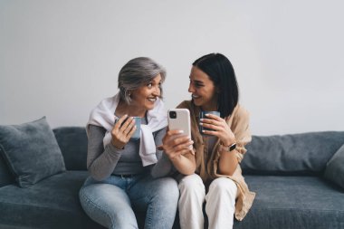 Mutlu genç kız ve yaşlı anne gündelik kıyafetleriyle akıllı telefon kullanıyor sosyal medyada sohbet ediyor ve gülüşüyor kanepede dinlenip içki içerken birbirlerine bakıyorlar.